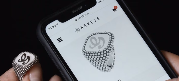 A dotted signet ring with a letter monogram held next to a phone showing the same ring in the Nove25 configurator on nove25.net.