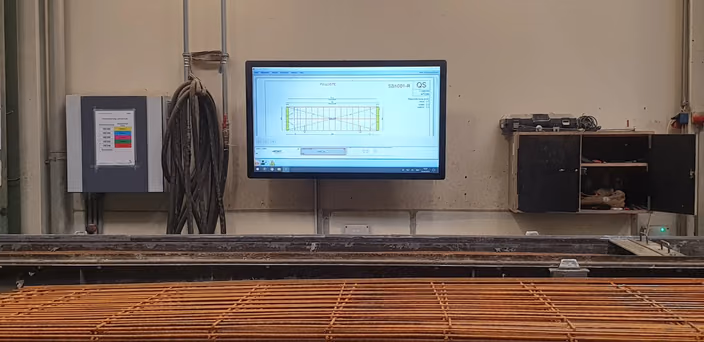 Shop floor view showing a monitor displaying a CAD blueprint for a concrete element, with a printed reference drawing on the wall.