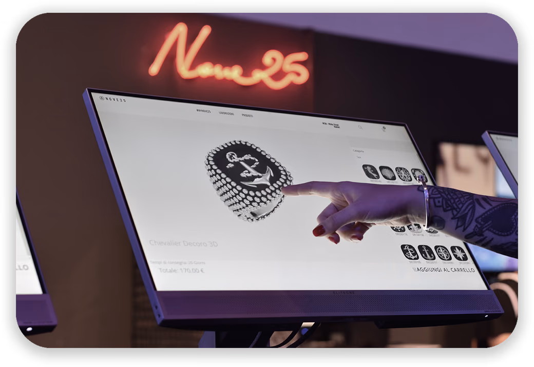 A hand with painted nails and a tattooed arm points at a signet ring with an anchor in the Nove25 configurator on an in-store touch screen, under a neon Nove25 sign.